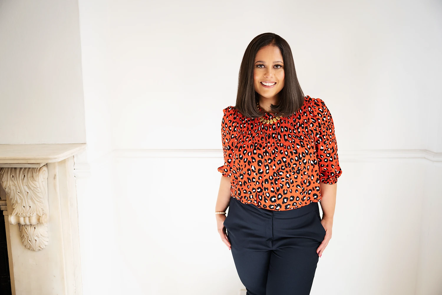 Caron Pollard, smiling, in a red leopard print top and navy trousers against a white wall